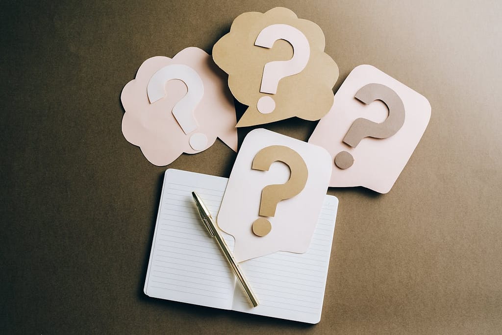 An open notebook with a pen lies on a desk, surrounded by four paper cutouts shaped like speech bubbles with question marks—suggesting the many questions surrounding lemon law vs warranty.