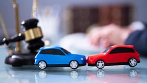 Two toy cars, one blue and one red, are positioned nose-to-nose on a table, with a law firm’s judge’s gavel and legal scales visible in the background.