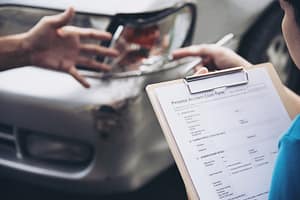 A person holds a clipboard with a personal accident claim form near a damaged car, while another person gestures toward the car’s dented front bumper.