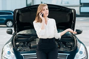 A woman in business attire sits on the front of a car with the hood open, talking on her cellphone and gesturing with her hand, appearing concerned, in an outdoor setting.