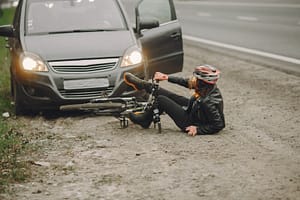 A person wearing a helmet and black clothes sits on the ground next to a fallen bicycle in front of a grey car with its door open, near the edge of a road.