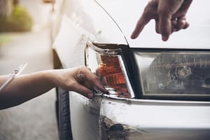 Two people examine and point at damage on the front bumper and headlight area of a white car, which shows visible scratches and dents.