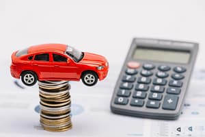 A red toy car sits on a tall stack of assorted coins, with a calculator and financial documents in the background, representing car expenses or auto finance calculations.