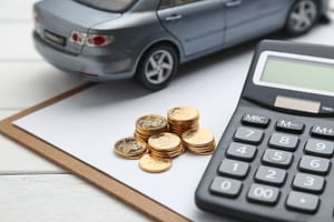 A clipboard with a blank sheet holds stacked coins, a black calculator, and a model car on a white table, suggesting car expenses or financial planning related to vehicles.