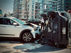 A white SUV with front-end damage is stopped next to a black vehicle overturned onto its side on a city street, with buildings and people in the background—a scene that highlights what to do after a car accident.