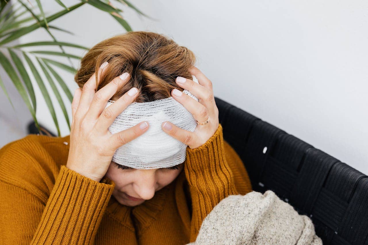 A person wearing a brown sweater sits with a bandaged head, holding their head in both hands, appearing to be in pain or discomfort—perhaps considering whether to file a personal injury claim.
