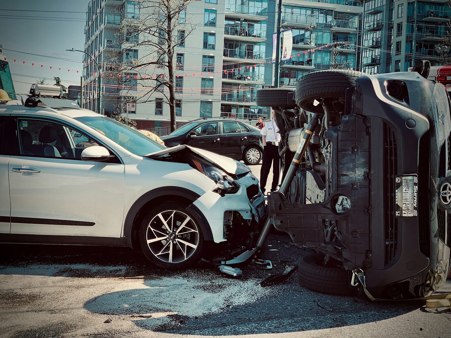 A white SUV with front-end damage is stopped next to a black vehicle overturned onto its side on a city street, with buildings and people in the background—a scene that highlights what to do after a car accident.