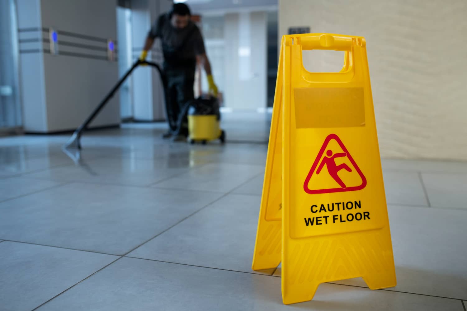 Caution Wet Floor sign placed in a tiled hallway while a cleaner works in the background
