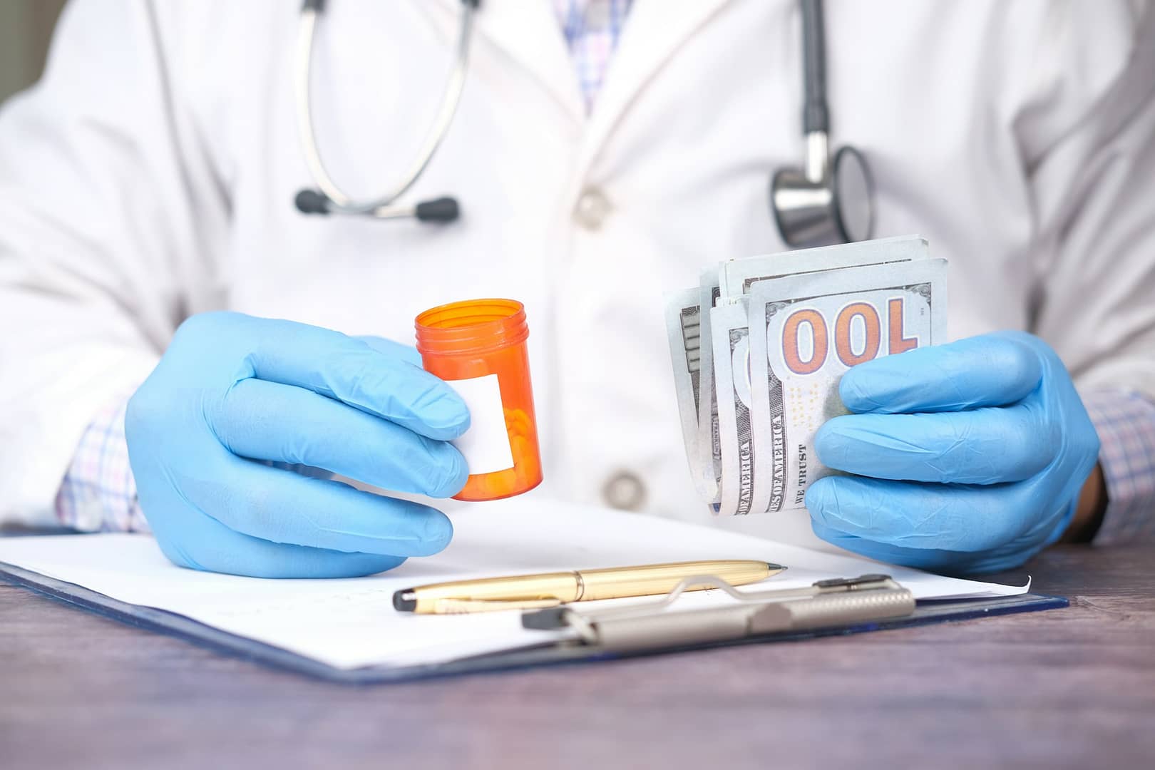 A person in a white coat and blue gloves holds a pill bottle and a stack of hundred-dollar bills, suggesting the rising cost of medical bills, with a stethoscope and paperwork on the desk.