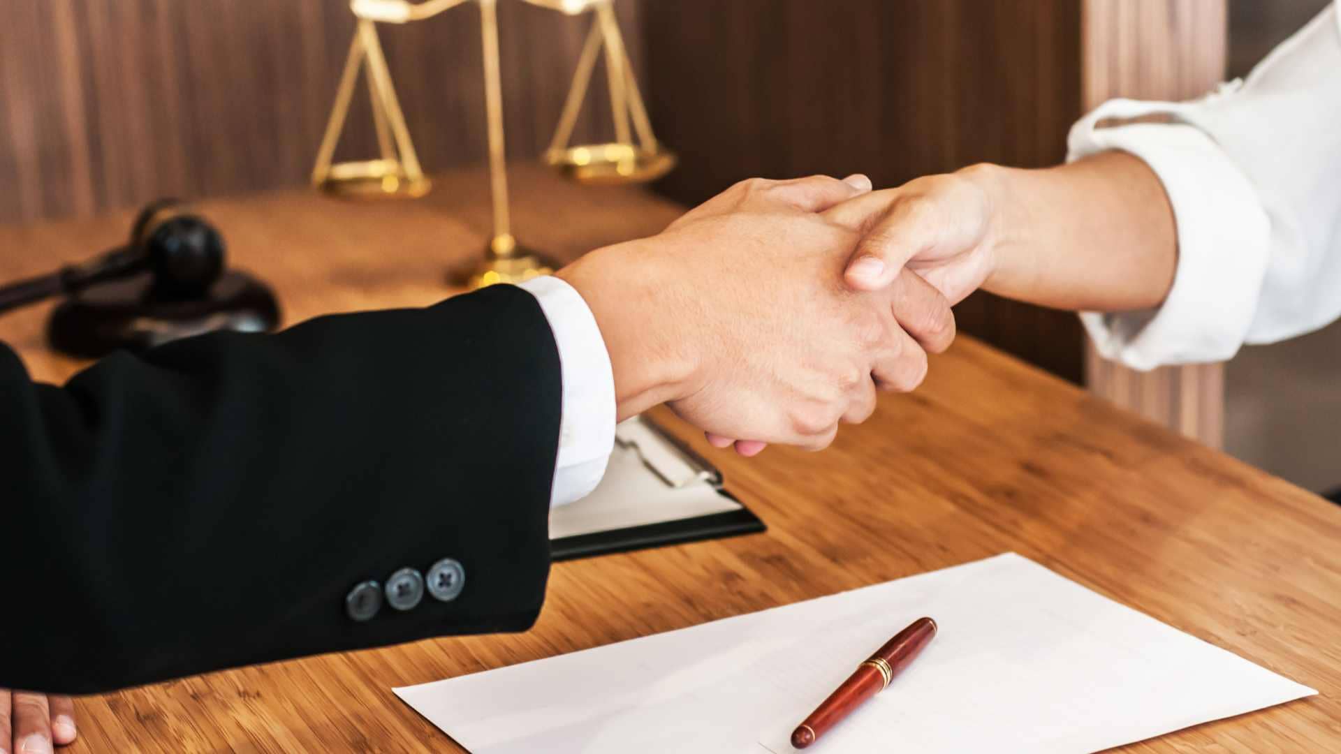 Two people shake hands across a wooden desk with a contract, pen, gavel, and scales of justice visible in the background, symbolizing the trusted guidance of California legal advocates.