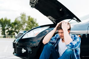Frustrated man sitting by a broken-down car with hood open, showing vehicle trouble