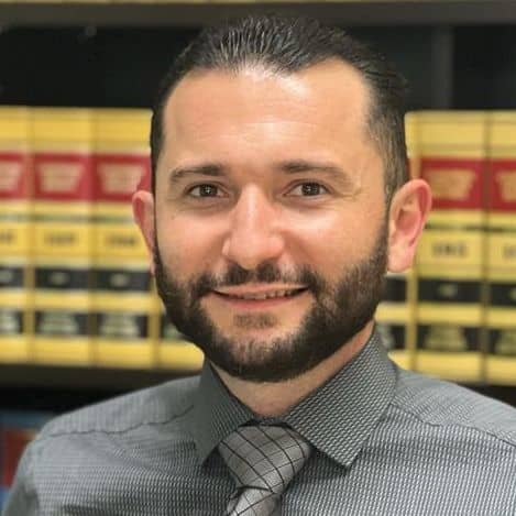 George Mkrtchyan, a man with short dark hair and a beard, wearing a collared shirt and tie, stands in front of shelves filled with legal books.